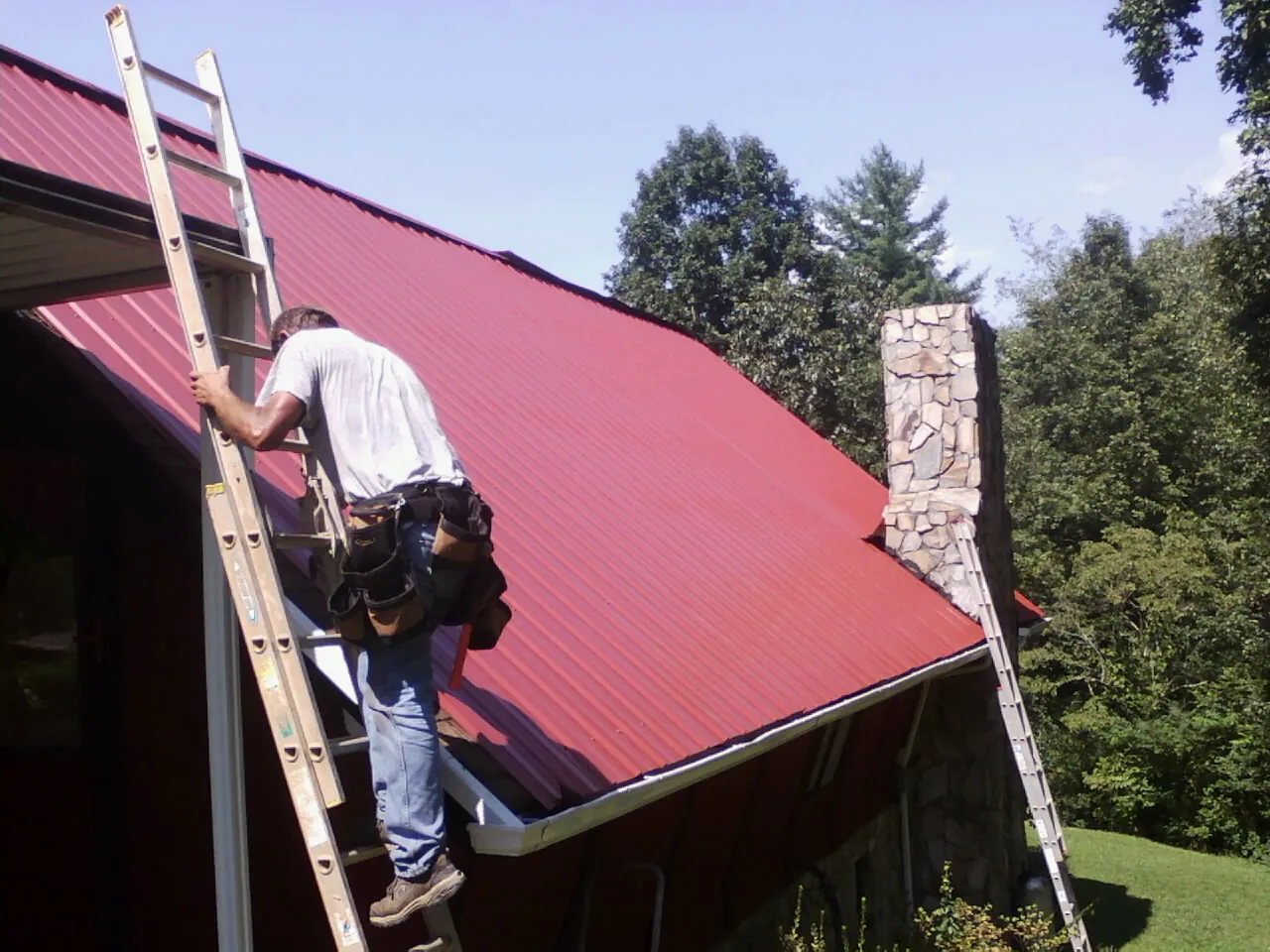 Roofer on a ladder beside a red metal roof and stone chimney — Soffit & Fascia Repair by Roofing Brothers in Demarest
